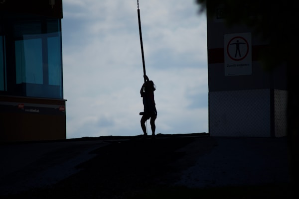 A silhouette of a person is in the center, swinging or holding onto a rope. The background is a slightly cloudy sky, providing contrast to the dark figure. There are structures on either side, one with a window and the other with a sign featuring a prohibited entry symbol.