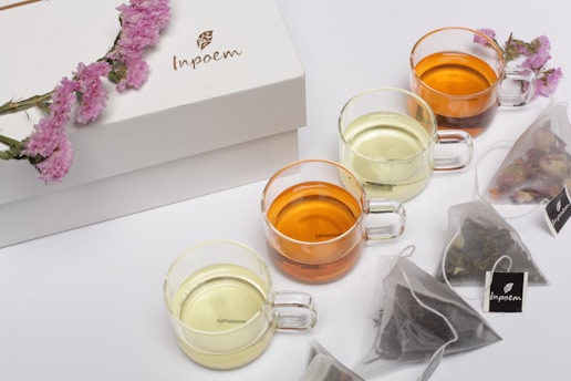 A set of four glass mugs filled with different shades of tea are arranged on a surface alongside transparent tea bags. A decorative white box and a sprig of pink flowers accompany the setup.