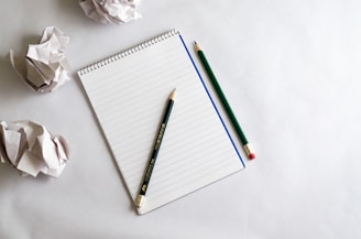A spiral-bound notebook lies on a white surface, accompanied by two green pencils. Several crumpled pieces of paper are scattered around, suggesting a creative or brainstorming environment.