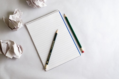A spiral-bound notebook lies on a white surface, accompanied by two green pencils. Several crumpled pieces of paper are scattered around, suggesting a creative or brainstorming environment.