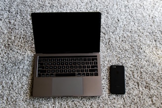 A laptop with a closed screen sitting on a light gray carpet, next to a black smartphone. The laptop is silver with a QWERTY keyboard, and the smartphone is positioned slightly below and to the right of the laptop.