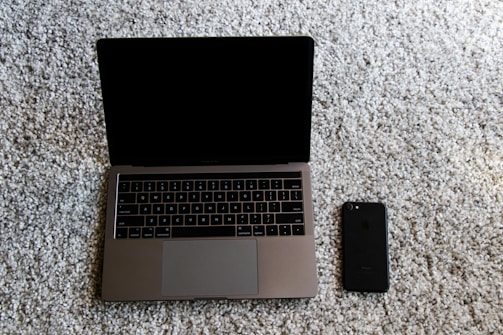 A laptop with a closed screen sitting on a light gray carpet, next to a black smartphone. The laptop is silver with a QWERTY keyboard, and the smartphone is positioned slightly below and to the right of the laptop.