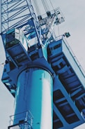 A large, industrial crane with metal components and a cabin for the operator is shown from a low angle. The machinery appears sturdy, with intricate details of its structure, including cables and railings that are prominently visible. The image has an industrial, mechanical feel.