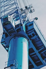 A large, industrial crane with metal components and a cabin for the operator is shown from a low angle. The machinery appears sturdy, with intricate details of its structure, including cables and railings that are prominently visible. The image has an industrial, mechanical feel.