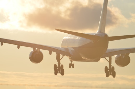 A large commercial airplane is in the process of landing or taking off, with its landing gear extended. The aircraft is backlit by a warm, golden sunset sky, creating a dramatic silhouette effect. Clouds are visible in the background, adding texture to the scene.