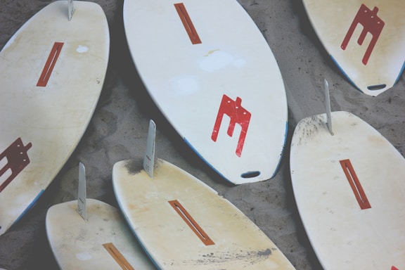Multiple surfboards are lying on a sandy surface, each with a distinct red design and single fin. The boards exhibit signs of use and vary slightly in color.