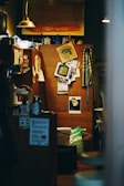 An interior space with a warm, cozy atmosphere features an eclectic arrangement of personal items. A bulletin board showcases various art pieces, photographs, and notes. Dim lighting is provided by small pendant lamps. A painting with warm tones hangs above, and various household items, including mugs and containers, are scattered around.