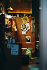An interior space with a warm, cozy atmosphere features an eclectic arrangement of personal items. A bulletin board showcases various art pieces, photographs, and notes. Dim lighting is provided by small pendant lamps. A painting with warm tones hangs above, and various household items, including mugs and containers, are scattered around.