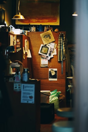 An interior space with a warm, cozy atmosphere features an eclectic arrangement of personal items. A bulletin board showcases various art pieces, photographs, and notes. Dim lighting is provided by small pendant lamps. A painting with warm tones hangs above, and various household items, including mugs and containers, are scattered around.