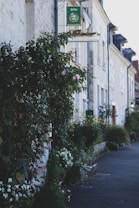 A charming, narrow street lined with traditional European-style buildings, featuring a dense display of greenery and blooming flowers along the walls. A sign for a g&icirc;te is visible above, indicating accommodations typical in France. The street has a peaceful, quaint atmosphere.