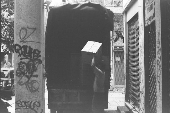 A person is lifting a large cardboard box onto a truck in a narrow alleyway. The truck is darkly covered, and the scene includes graffiti on the walls and a partially visible street. The setting is urban and captures an everyday moment.