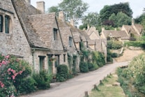 A quaint stone street lined with traditional English cottages featuring steeply pitched roofs. Vibrant green foliage and colorful flowers adorn the scenes, while a narrow path runs alongside a small stream. The architecture reflects historical charm with neatly trimmed hedges and well-maintained gardens.