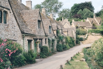 A quaint stone street lined with traditional English cottages featuring steeply pitched roofs. Vibrant green foliage and colorful flowers adorn the scenes, while a narrow path runs alongside a small stream. The architecture reflects historical charm with neatly trimmed hedges and well-maintained gardens.