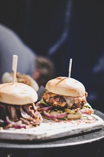 Two gourmet burgers are presented on a wooden board, each topped with unique ingredients. The burger on the right features a succulent patty with melted cheese, onions, and leafy greens. The burger on the left has a shredded meat topping, possibly with a barbecue sauce. Both are served in soft buns with a stick inserted to hold them together.