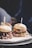 Two gourmet burgers are presented on a wooden board, each topped with unique ingredients. The burger on the right features a succulent patty with melted cheese, onions, and leafy greens. The burger on the left has a shredded meat topping, possibly with a barbecue sauce. Both are served in soft buns with a stick inserted to hold them together.