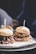 Two gourmet burgers are presented on a wooden board, each topped with unique ingredients. The burger on the right features a succulent patty with melted cheese, onions, and leafy greens. The burger on the left has a shredded meat topping, possibly with a barbecue sauce. Both are served in soft buns with a stick inserted to hold them together.