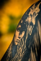 A black and white tattoo is depicted on skin, featuring a detailed face with long, flowing hair. Above the face, a skull is intricately integrated into the design, creating a mysterious and artistic composition. The background is a blurred gradient of yellow and green, enhancing the contrast of the tattoo art.