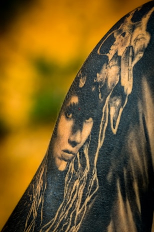 A black and white tattoo is depicted on skin, featuring a detailed face with long, flowing hair. Above the face, a skull is intricately integrated into the design, creating a mysterious and artistic composition. The background is a blurred gradient of yellow and green, enhancing the contrast of the tattoo art.