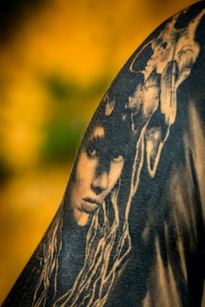 A black and white tattoo is depicted on skin, featuring a detailed face with long, flowing hair. Above the face, a skull is intricately integrated into the design, creating a mysterious and artistic composition. The background is a blurred gradient of yellow and green, enhancing the contrast of the tattoo art.