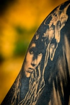 A black and white tattoo is depicted on skin, featuring a detailed face with long, flowing hair. Above the face, a skull is intricately integrated into the design, creating a mysterious and artistic composition. The background is a blurred gradient of yellow and green, enhancing the contrast of the tattoo art.