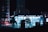 A night-time urban scene showcasing a well-lit modern building in the city center with large illuminated signage reading 'Bahnhof Potsdamer Platz'. The building features extensive glass windows that reflect a cool blue glow. In the foreground, silhouettes of trees and bicycles are visible, while tall city buildings with lit windows stand in the background.