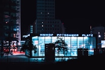 A night-time urban scene showcasing a well-lit modern building in the city center with large illuminated signage reading 'Bahnhof Potsdamer Platz'. The building features extensive glass windows that reflect a cool blue glow. In the foreground, silhouettes of trees and bicycles are visible, while tall city buildings with lit windows stand in the background.