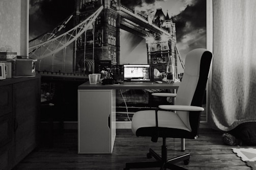 A monochrome photograph of a home office setting featuring a desk with a laptop open, accompanied by a water bottle and glass. The chair is positioned in front of the desk, and a large mural of a famous bridge is on the wall behind the setup.