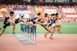 A group of athletes is competing in a hurdle race on a track. Each runner is captured mid-air as they leap over barriers in a stadium setting. The background includes red stadium seating and various banners. The athletes wear numbered jerseys and running shoes.
