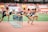 A group of athletes is competing in a hurdle race on a track. Each runner is captured mid-air as they leap over barriers in a stadium setting. The background includes red stadium seating and various banners. The athletes wear numbered jerseys and running shoes.