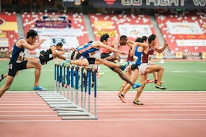 A group of athletes is competing in a hurdle race on a track. Each runner is captured mid-air as they leap over barriers in a stadium setting. The background includes red stadium seating and various banners. The athletes wear numbered jerseys and running shoes.