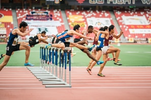 A group of athletes is competing in a hurdle race on a track. Each runner is captured mid-air as they leap over barriers in a stadium setting. The background includes red stadium seating and various banners. The athletes wear numbered jerseys and running shoes.