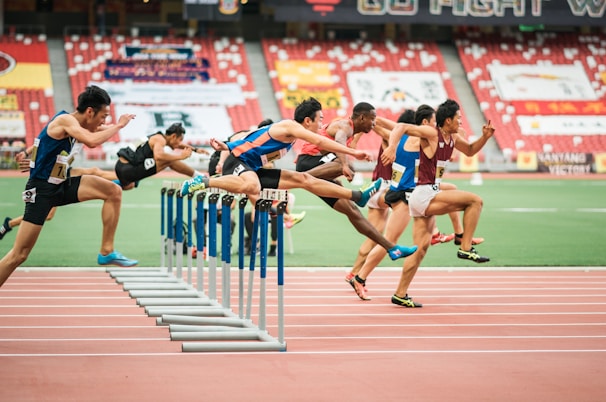 A group of athletes is competing in a hurdle race on a track. Each runner is captured mid-air as they leap over barriers in a stadium setting. The background includes red stadium seating and various banners. The athletes wear numbered jerseys and running shoes.