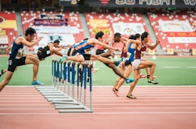A group of athletes is competing in a hurdle race on a track. Each runner is captured mid-air as they leap over barriers in a stadium setting. The background includes red stadium seating and various banners. The athletes wear numbered jerseys and running shoes.