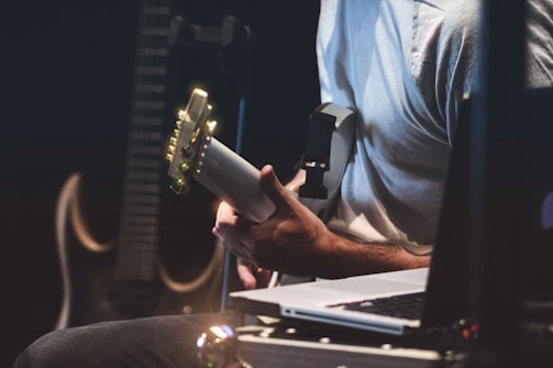 A person is playing an electric guitar, surrounded by music equipment such as a laptop and another guitar in the background. The setting is dimly lit, possibly suggesting a studio or performance environment.