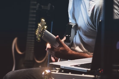A person is playing an electric guitar, surrounded by music equipment such as a laptop and another guitar in the background. The setting is dimly lit, possibly suggesting a studio or performance environment.