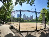 A baseball field enclosed by a black metal fence with a city skyline visible in the background. The field is surrounded by tall green trees and there is a set of bleachers to the left. The sky is clear and blue, with sunlight casting dappled shadows on the ground.