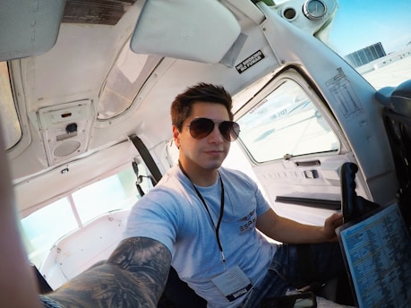 A person with sunglasses is seated inside a small airplane cockpit. The individual is wearing a light gray T-shirt and has a tattoo sleeve on their left arm. The cockpit interior is predominantly white with various controls and a 'NO SMOKING' sign visible. The person is holding a checklist or document in their right hand.