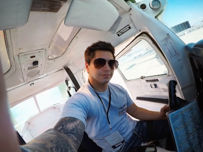 A person with sunglasses is seated inside a small airplane cockpit. The individual is wearing a light gray T-shirt and has a tattoo sleeve on their left arm. The cockpit interior is predominantly white with various controls and a 'NO SMOKING' sign visible. The person is holding a checklist or document in their right hand.