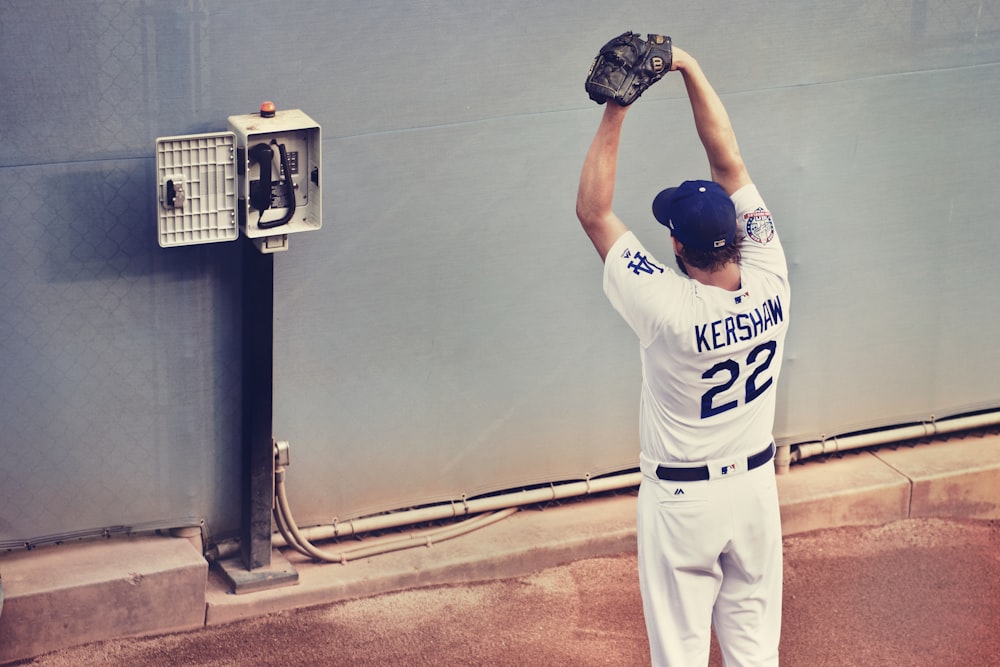 man in white baseball jersey shirt standing with gloves in front of telephone on wall