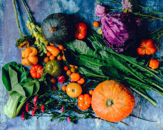 A vibrant assortment of fresh vegetables and flowers arranged on a textured blue surface. The selection includes an orange pumpkin, a green kabocha squash, various orange and red bell peppers, cherry tomatoes, purple cabbage, and an array of green leafy vegetables. Among the vegetables are also blooms of sunflowers and pink flowers, adding a touch of floral contrast to the colorful spread.