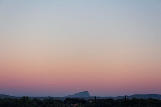 A tranquil landscape with a vast sky transitioning through soft pastel hues of pink, orange, and blue, casting a serene glow over distant mountains and a lush green horizon.