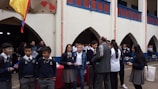A group of students and several adults are gathered in a school courtyard. Some students are in uniform, and there are decorative elements like red hearts on the walls. A yellow, blue, and red flag is visible. A few people appear to be holding papers or clipboards.