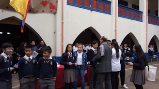 A group of students and several adults are gathered in a school courtyard. Some students are in uniform, and there are decorative elements like red hearts on the walls. A yellow, blue, and red flag is visible. A few people appear to be holding papers or clipboards.