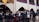 A group of students and several adults are gathered in a school courtyard. Some students are in uniform, and there are decorative elements like red hearts on the walls. A yellow, blue, and red flag is visible. A few people appear to be holding papers or clipboards.