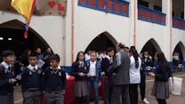 A group of students and several adults are gathered in a school courtyard. Some students are in uniform, and there are decorative elements like red hearts on the walls. A yellow, blue, and red flag is visible. A few people appear to be holding papers or clipboards.