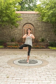 A woman is practicing a yoga pose, standing on one leg with hands in a praying position. She is in an outdoor setting with lush green trees surrounding a brick wall and a circular stone pattern on the ground.
