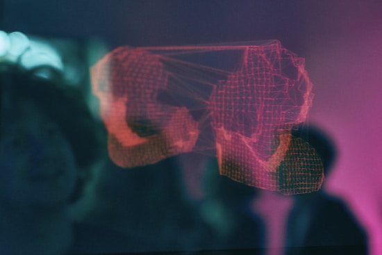 A digital wireframe of boxing gloves illuminated in red appears against a blurred, dark background. The design seems to be part of a 3D display or hologram, projecting a modern and futuristic aesthetic. Nearby, there is an indistinct silhouette of a person observing the display.