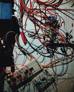A complex tangle of multicolored wires is connected to several electronic breadboards and circuits, suggesting an environment of experimentation or electronic prototyping. The wires cover a significant portion of the wall space, intertwining in a chaotic but purposeful manner.