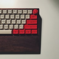 A mechanical keyboard with a wooden base featuring a combination of white and red keycaps. The white keycaps have Japanese characters on them, while the red keycaps are located on the right side of the keyboard and include various function keys. A red rose-shaped keycap can be seen in the top right corner.