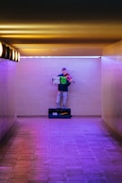 A person stands in an underground passage, playing a violin. They are positioned against a wall, with a music stand in front of them holding sheet music. The corridor is illuminated with warm yellow overhead lights on one side and purple lighting on the floor and lower part of the wall.